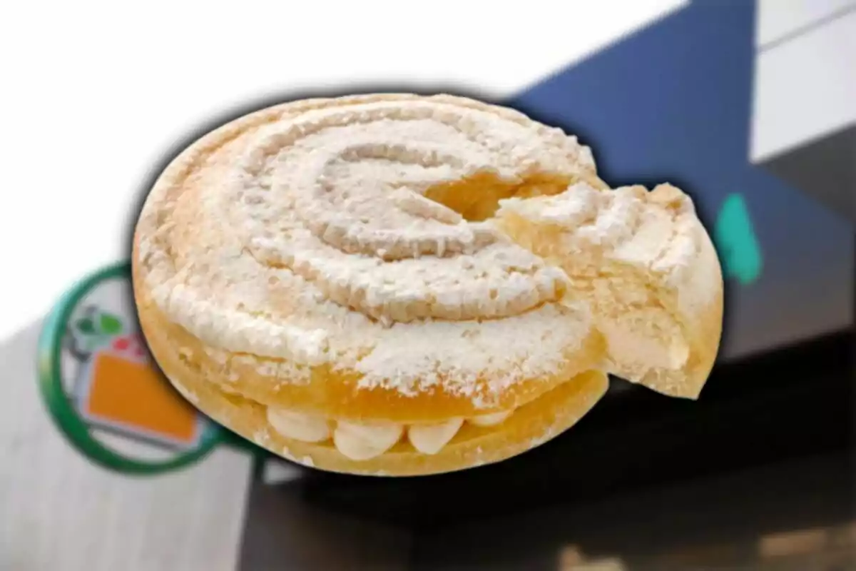 Un pa dolç tipus petxina farcit de crema amb una mossegada, al fons s’observa la façana d’una botiga. Un pa dolç tipus petxina farcit de crema amb una mossegada, al fons s’observa la façana d’una botiga.