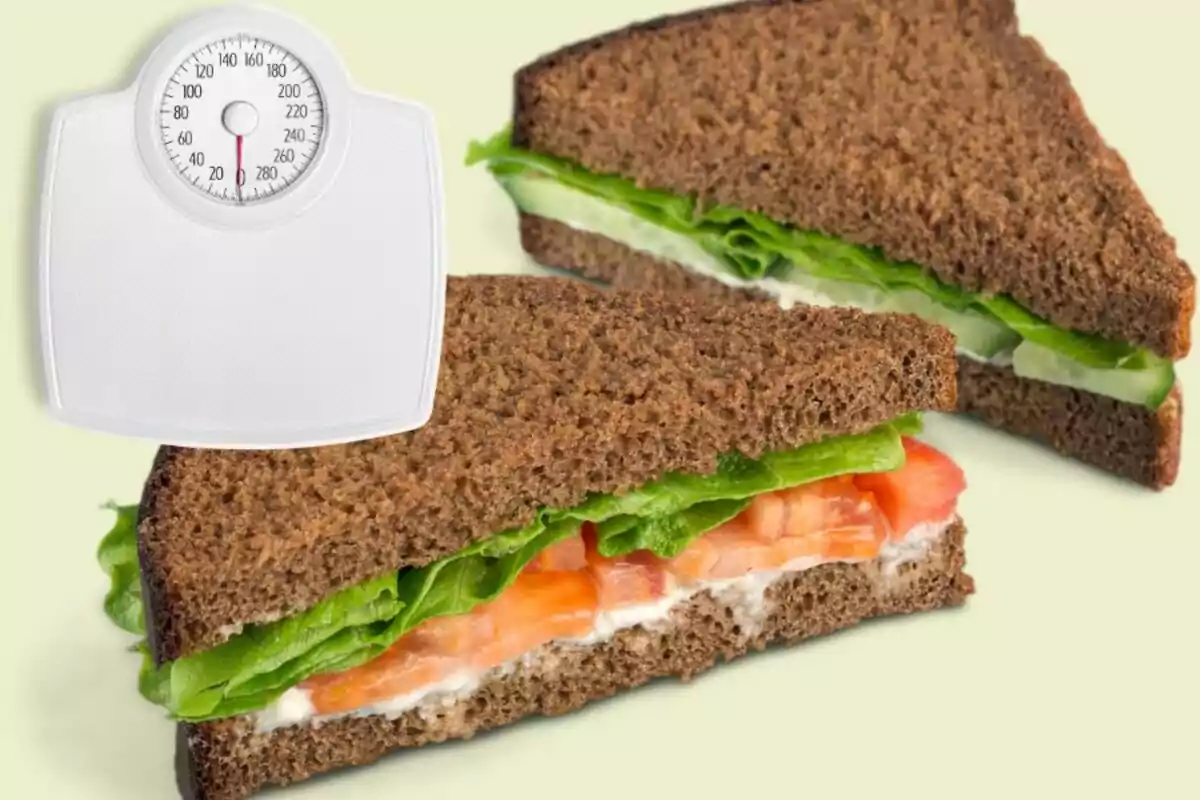 Sándwich de pan integral con lechuga y jitomate junto a una báscula blanca sobre fondo claro