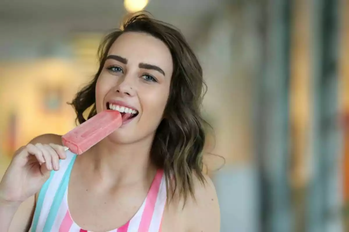 Dona jove somrient gaudint d’un gelat de paleta rosa en un ambient interior il·luminat Dona jove somrient gaudint d’un gelat de paleta rosa en un ambient interior il·luminat