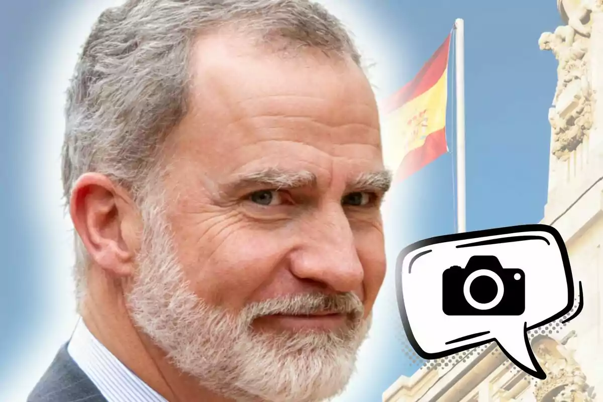 Primer pla del rei Felip de Borbó somrient amb una bandera d’Espanya onejant al fons i una icona de càmera a la cantonada inferior dreta.