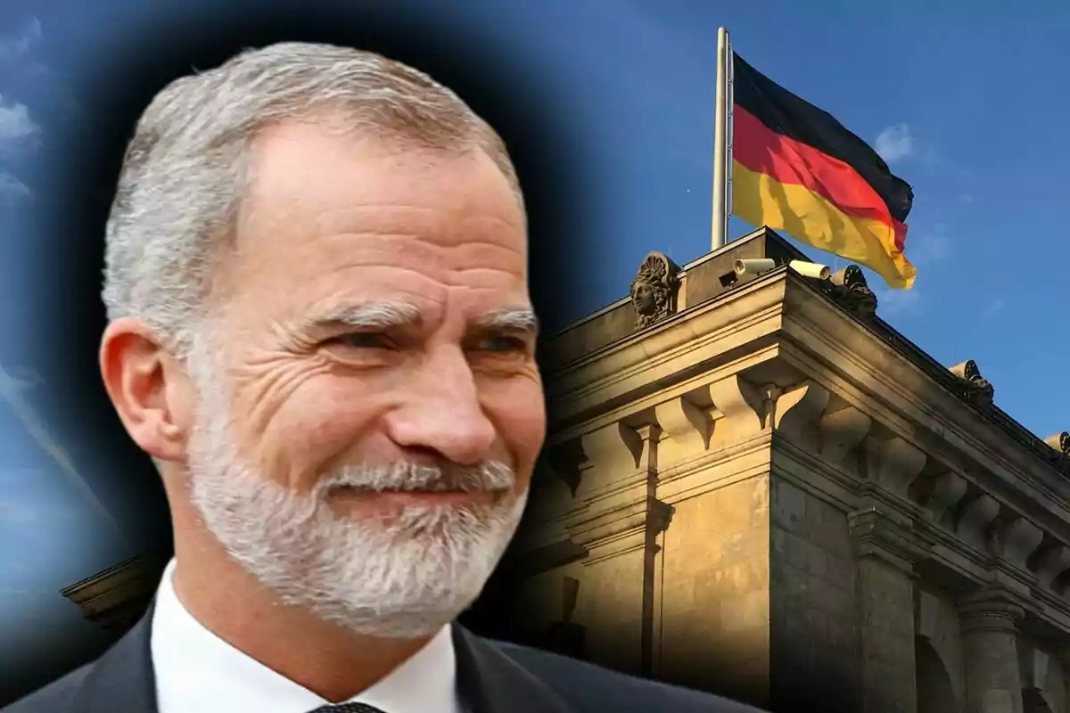 El rei Felip VI somrient en primer pla amb un edifici històric i una bandera d'Alemanya onejant al fons.