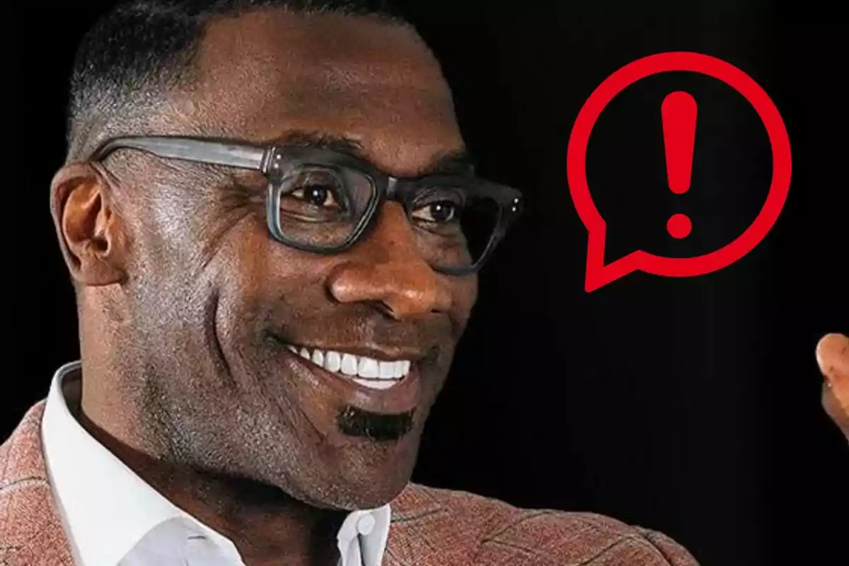 Shannon Sharpe sonriente con gafas y saco claro al lado de un ícono de advertencia en forma de globo de diálogo rojo sobre fondo negro