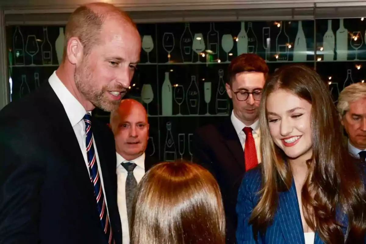 El príncipe Guillermo y la princesa Leonor sonríen mientras conversan con una niña de espaldas en un evento formal, rodeados de otras personas elegantemente vestidas.