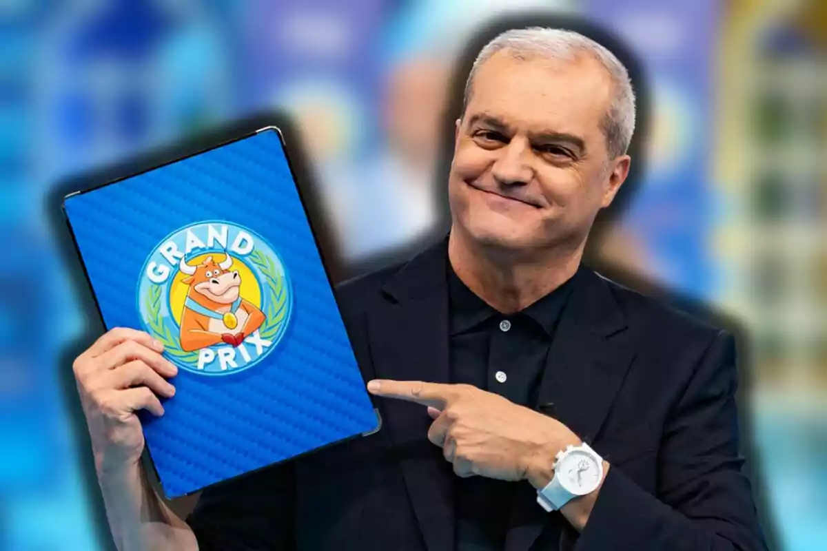 Ramón García sonriente con cabello canoso sostiene una carpeta azul con el logo de Grand Prix y una vaca caricaturizada mientras señala el logo