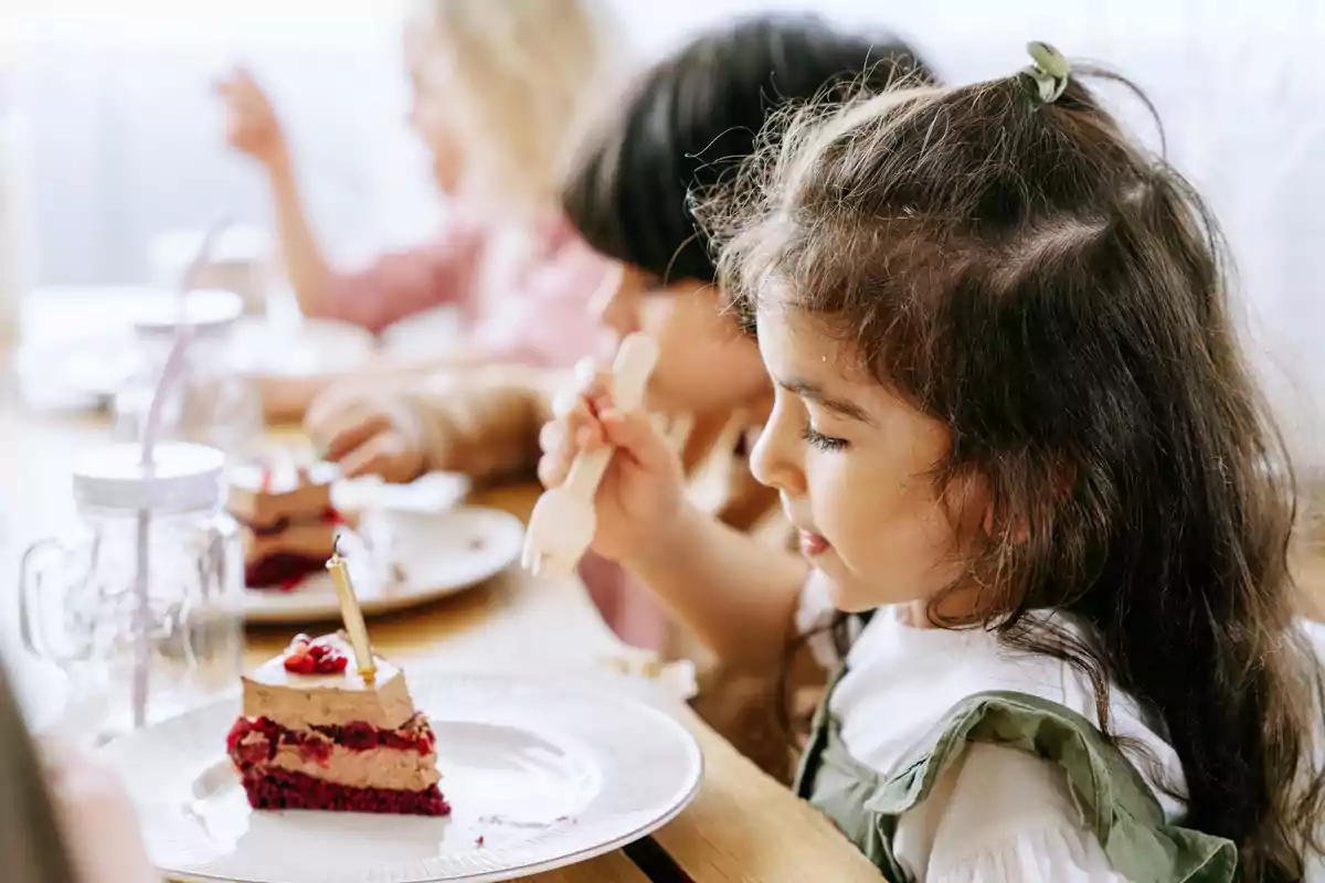 Little girl sitting at the table enjoying a piece of cake with other children in the background Little girl sitting at the table enjoying a piece of cake with other children in the background