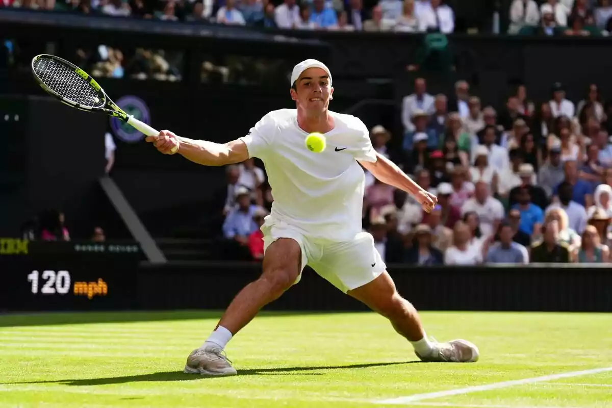 Tennista vestit de blanc, que és l’Oliver Tarvet, colpejant una pilota durant un partit en una pista de gespa amb públic de fons.
