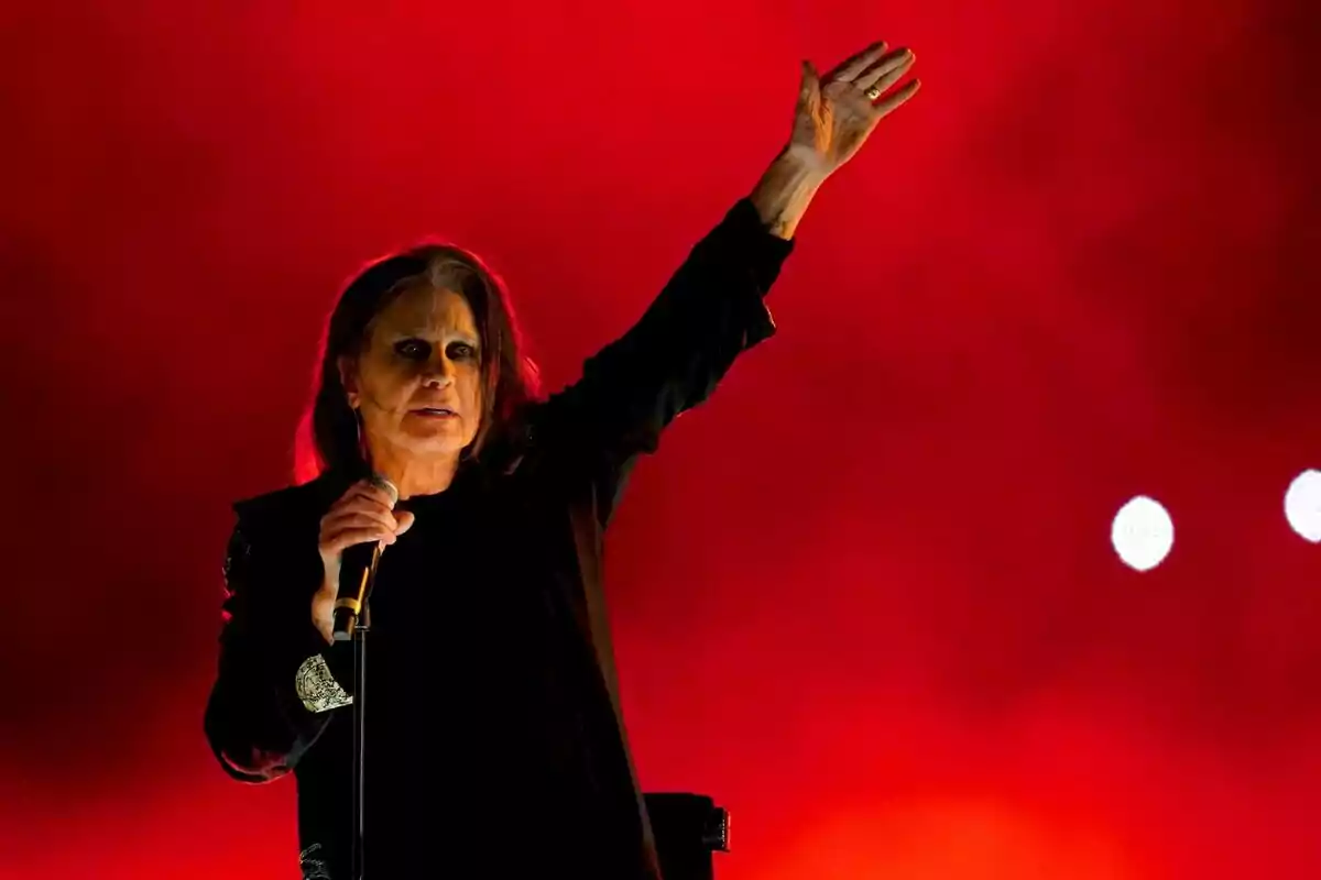 Ozzy Ousbourne con cabello largo y oscuro con ropa negra sosteniendo un micrófono y levantando una mano frente a un fondo rojo intenso Ozzy Ousbourne con cabello largo y oscuro con ropa negra sosteniendo un micrófono y levantando una mano frente a un fondo rojo intenso