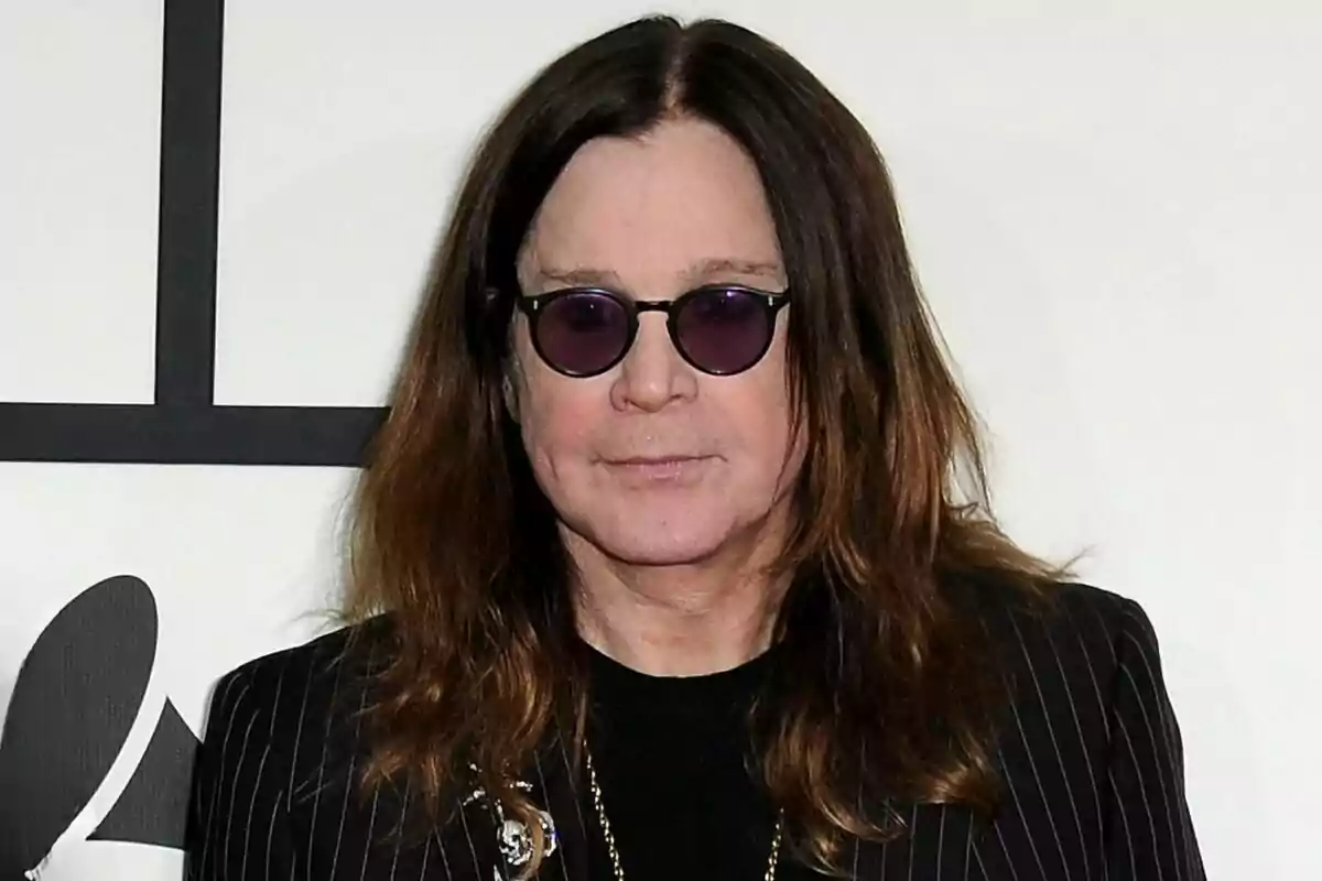 Ozzy Osbourne con cabello largo castaño, gafas de sol redondas y chaqueta de rayas posando frente a un fondo claro Ozzy Osbourne con cabello largo castaño, gafas de sol redondas y chaqueta de rayas posando frente a un fondo claro
