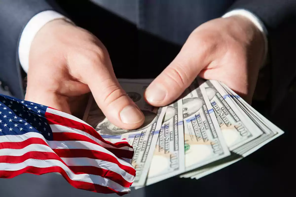 Hands holding hundred-dollar bills next to a United States flag Hands holding hundred-dollar bills next to a United States flag