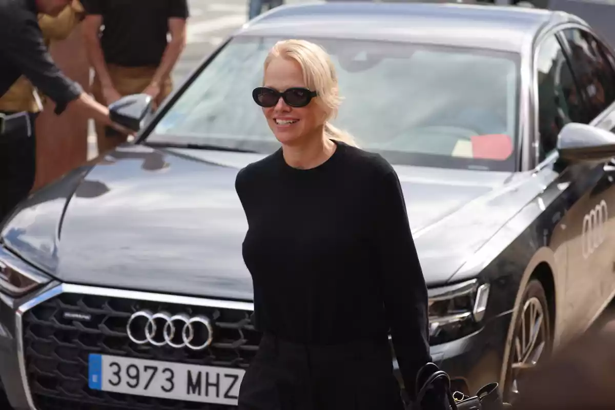 Pamela Anderson con gafas de sol y suéter negro sonríe mientras camina frente a un auto Audi plateado Pamela Anderson con gafas de sol y suéter negro sonríe mientras camina frente a un auto Audi plateado