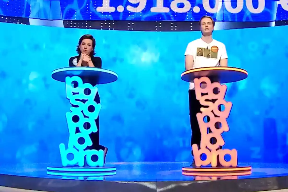 La Rosa i en Manu estan drets darrere de faristols de colors en un plató de televisió amb fons blau, participant en un concurs. Captura de 'Pasapalabra' La Rosa i en Manu estan drets darrere de faristols de colors en un plató de televisió amb fons blau, participant en un concurs. Captura de 'Pasapalabra'