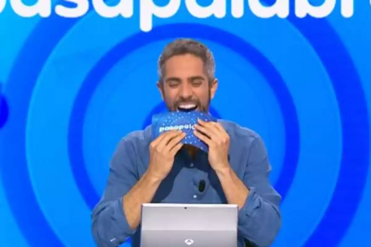 Roberto Leal somrient amb barba i camisa blava sosté una targeta blava de Pasapalabra davant de la seva boca en un plató de televisió amb fons blau. Roberto Leal somrient amb barba i camisa blava sosté una targeta blava de Pasapalabra davant de la seva boca en un plató de televisió amb fons blau.