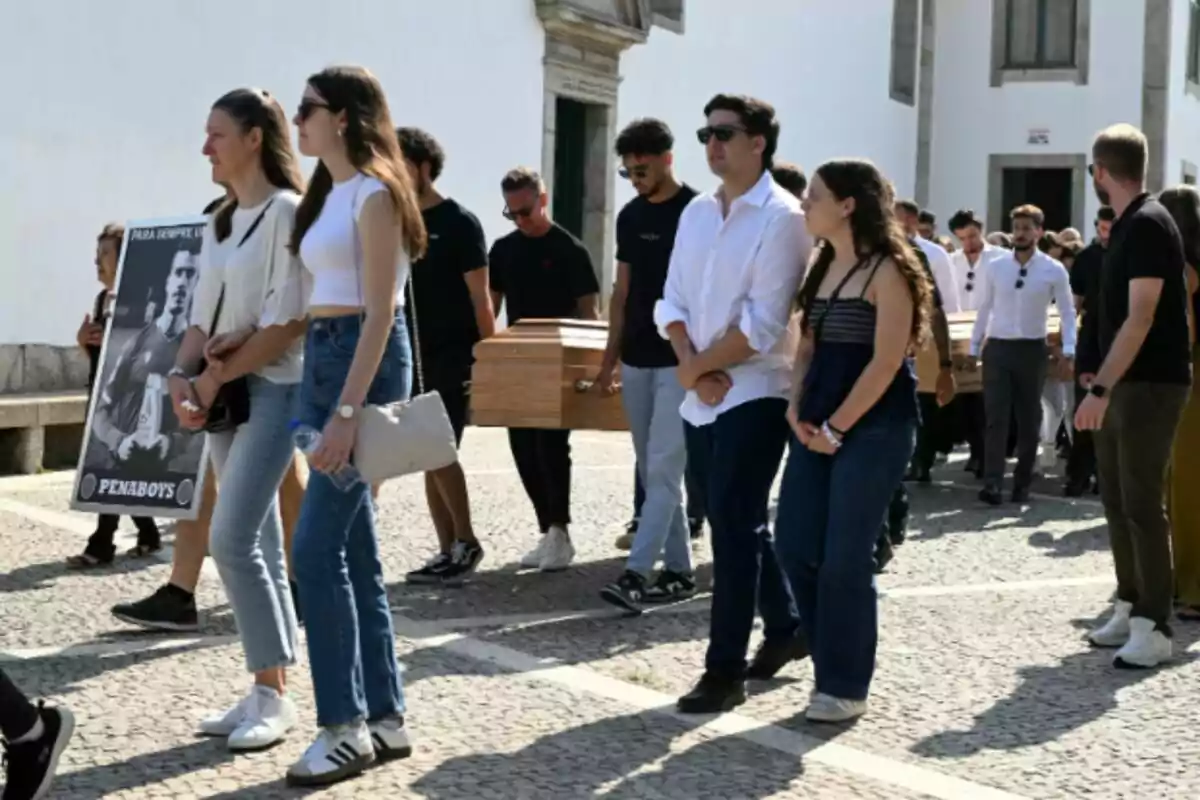 People walking in an outdoor funeral procession, some of them carrying a wooden coffin and others holding a sign with the image of a man, who is Diogo Jota. People walking in an outdoor funeral procession, some of them carrying a wooden coffin and others holding a sign with the image of a man, who is Diogo Jota.