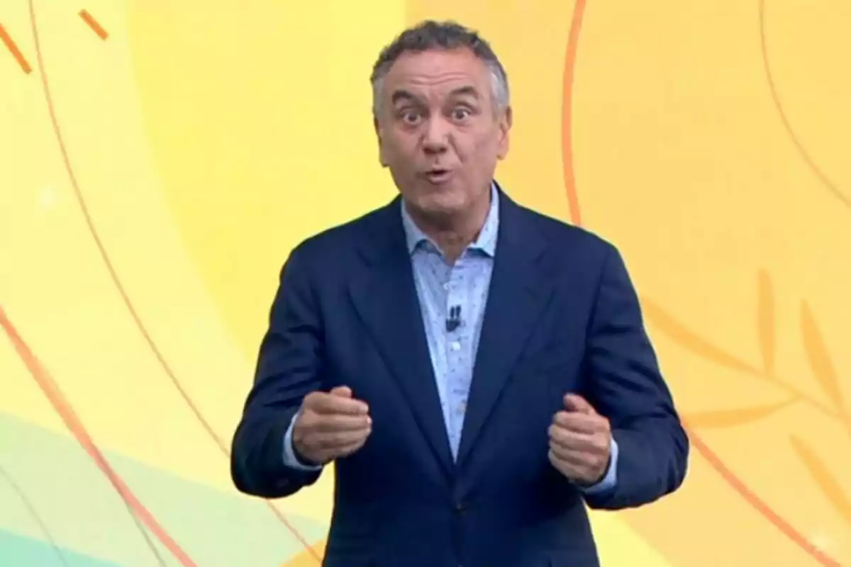 Pla mitjà de Roberto Brasero vestit amb un vestit blau i una camisa clara gesticulant davant d’un fons groc amb línies corbes. Pla mitjà de Roberto Brasero vestit amb un vestit blau i una camisa clara gesticulant davant d’un fons groc amb línies corbes.
