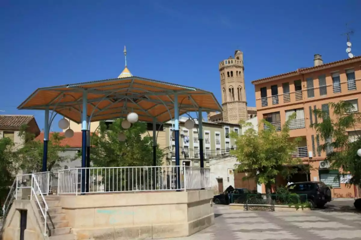 Quiosco en una plaza rodeada de árboles y edificios con una torre de ladrillo al fondo bajo un cielo despejado Quiosco en una plaza rodeada de árboles y edificios con una torre de ladrillo al fondo bajo un cielo despejado