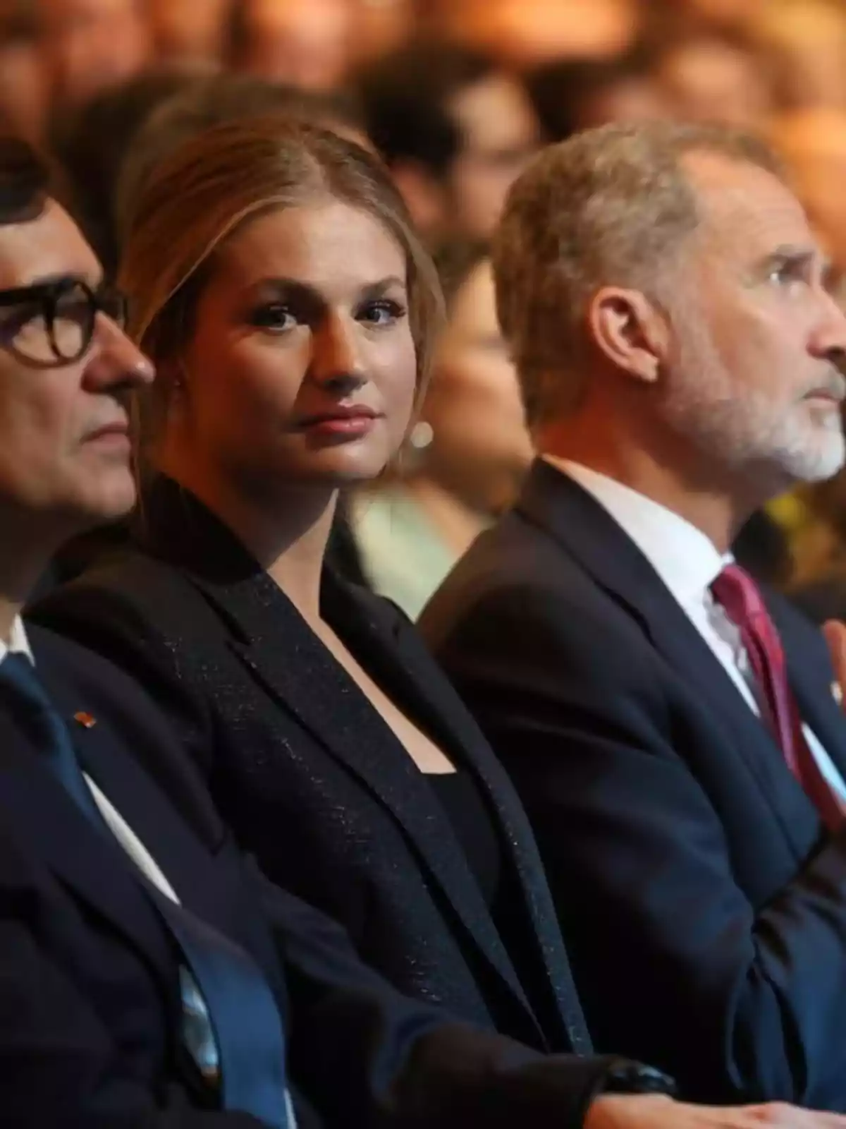 Salvador Illa, Princess Leonor, and Felipe VI seated at a formal event with the woman in the center looking at the camera and the men on either side facing forward.