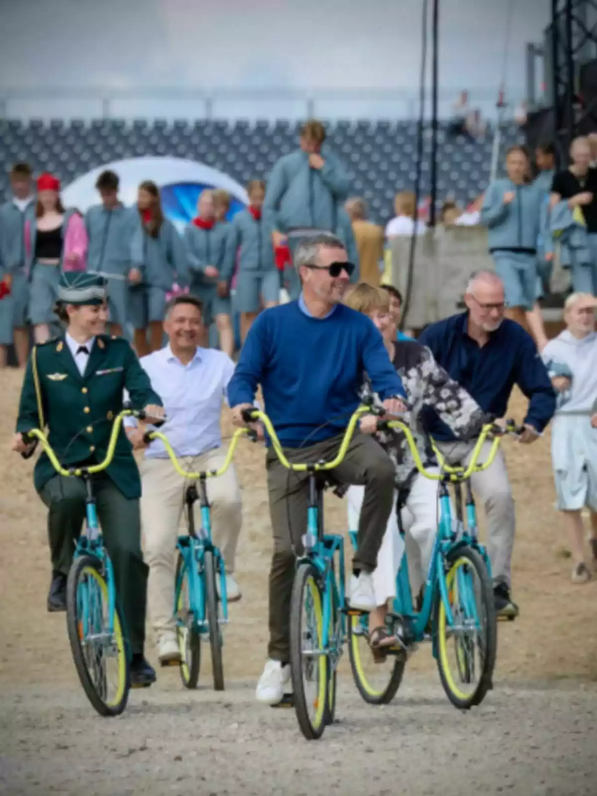 Group of people riding blue bicycles with yellow handlebars at an outdoor event while other people walk in the background Group of people riding blue bicycles with yellow handlebars at an outdoor event while other people walk in the background