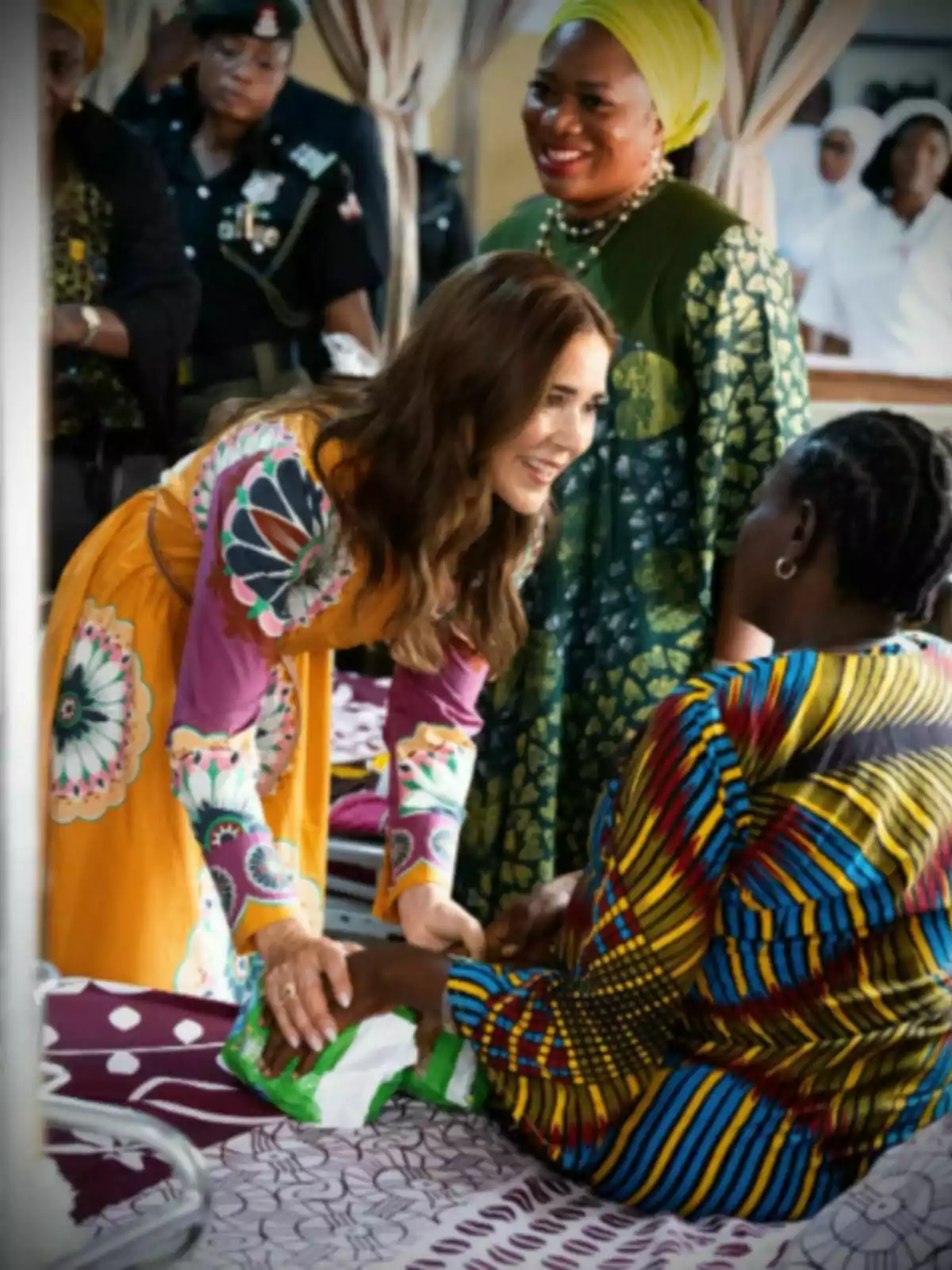 Mary of Denmark, wearing a colorful dress, smiles and holds the hand of another woman sitting on a bed while other people watch in an indoor setting. Mary of Denmark, wearing a colorful dress, smiles and holds the hand of another woman sitting on a bed while other people watch in an indoor setting.