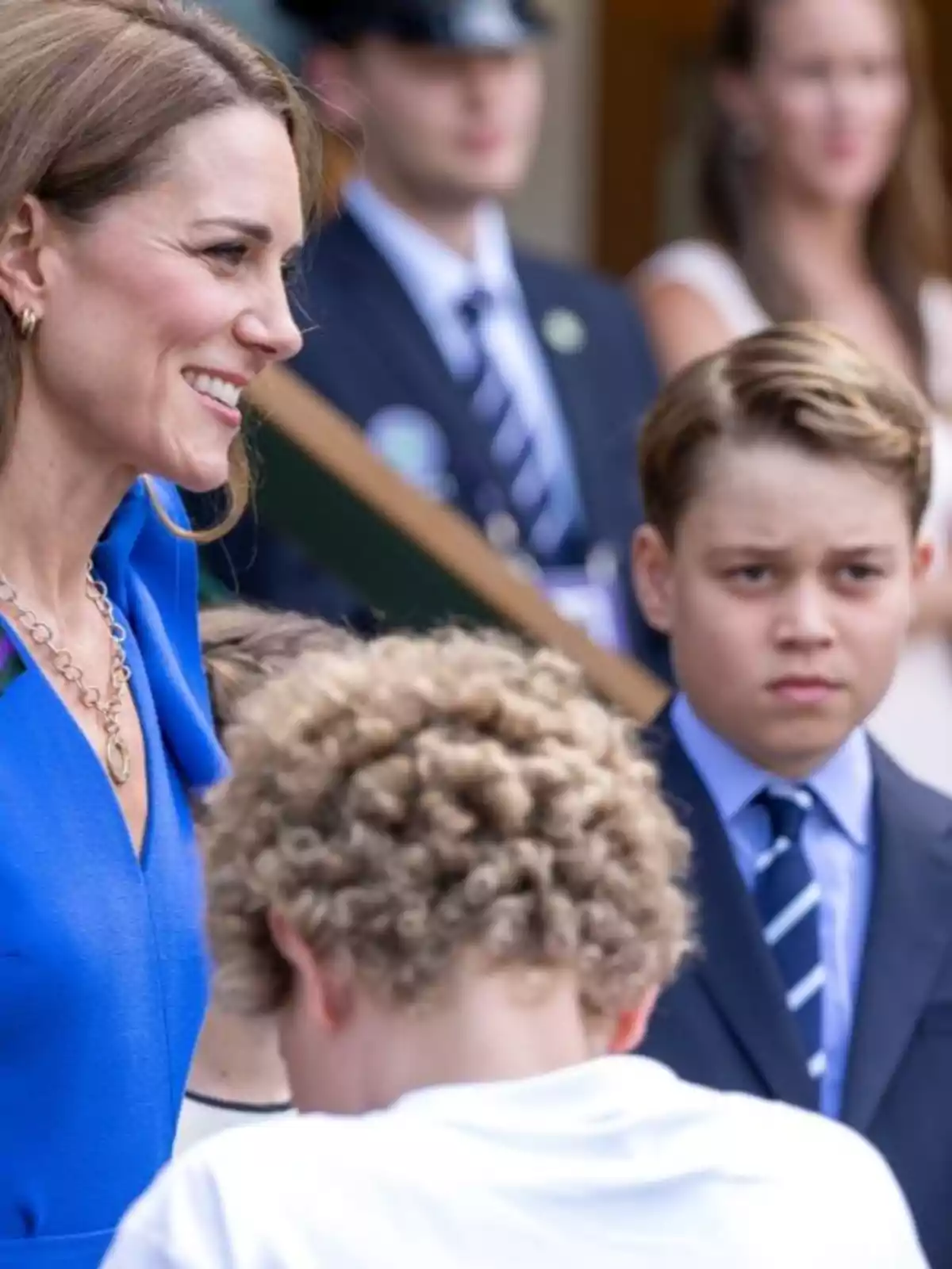 Kate Middleton somrient amb vestit blau està acompanyada pel príncep George amb vestit i corbata i un altre nen de cabells arrissats en primer pla, mentre diverses persones observen al fons. Kate Middleton somrient amb vestit blau està acompanyada pel príncep George amb vestit i corbata i un altre nen de cabells arrissats en primer pla, mentre diverses persones observen al fons.
