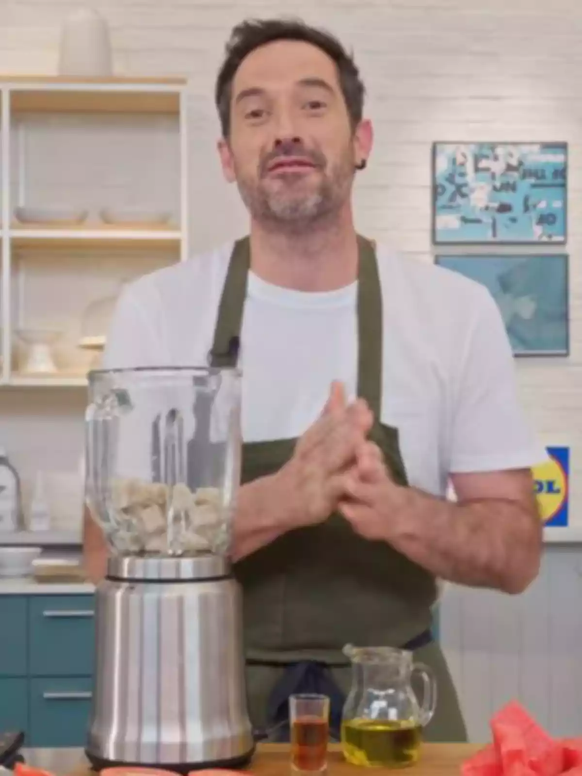 Joseba Arguiñano amb una samarreta blanca i un davantal verd és a una cuina davant d’una liquadora amb ingredients i recipients de vidre sobre la taula.