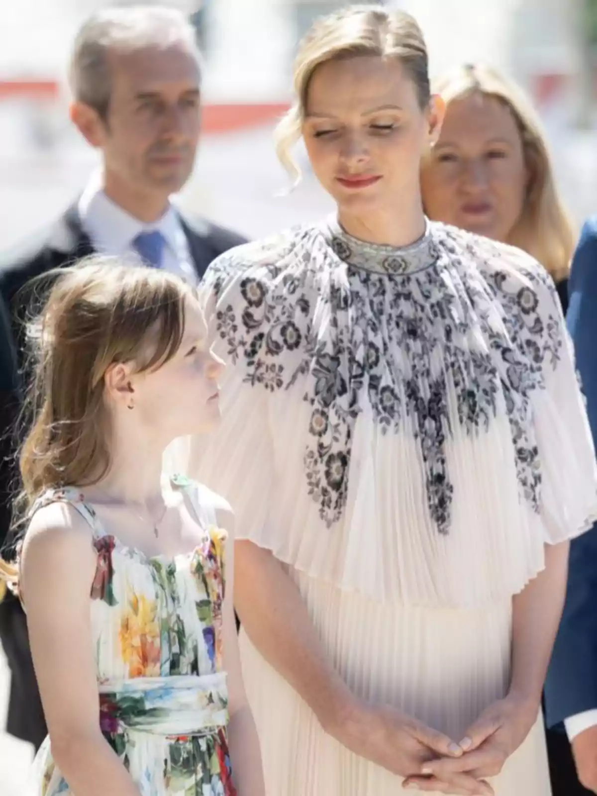 Charlene, amb un vestit blanc amb detalls florals en negre, mira amb tendresa a Gabriella, que porta un vestit de colors; ambdues són a l’aire lliure i hi ha persones desenfocades al fons. Charlene, amb un vestit blanc amb detalls florals en negre, mira amb tendresa a Gabriella, que porta un vestit de colors; ambdues són a l’aire lliure i hi ha persones desenfocades al fons.