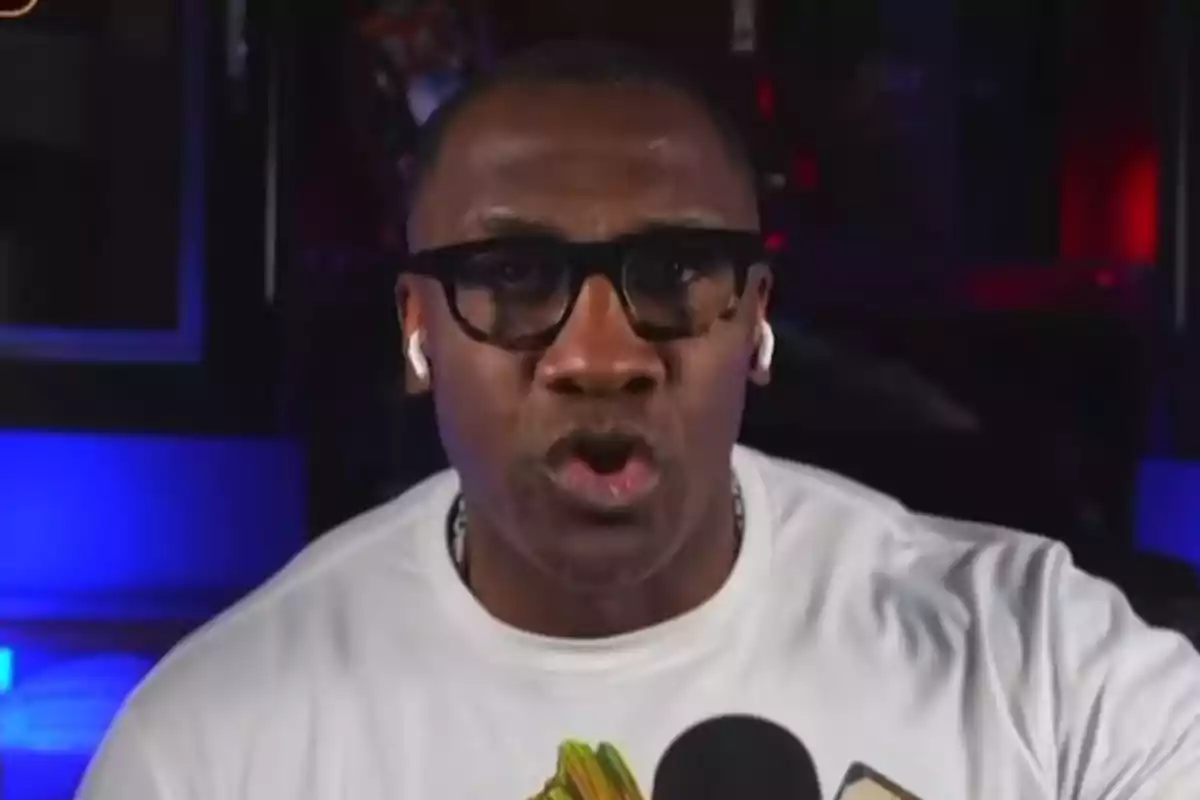 Shannon Sharpe con gafas y auriculares hablando frente a un micrófono en un ambiente con luces azules y rojas Shannon Sharpe con gafas y auriculares hablando frente a un micrófono en un ambiente con luces azules y rojas