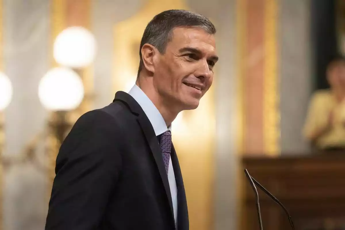 Home de cabell curt i canós amb vestit fosc i corbata, somrient mentre parla en un micròfon en un entorn elegant i formal