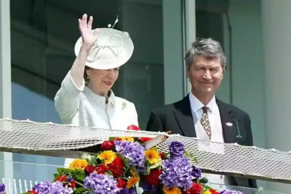 Una dona vestida de blanc amb barret saluda mentre està dreta al costat d’un home amb vestit fosc, tots dos darrere d’una barana decorada amb flors de colors. Una dona vestida de blanc amb barret saluda mentre està dreta al costat d’un home amb vestit fosc, tots dos darrere d’una barana decorada amb flors de colors.