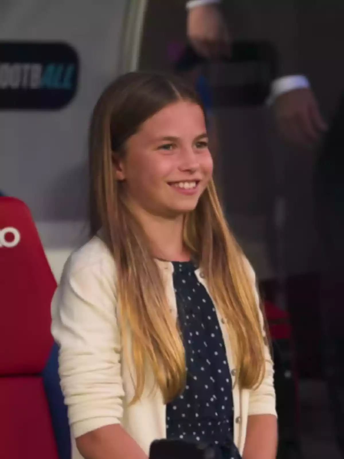 Long-haired blonde girl smiling while standing in a blue polka dot dress and a light sweater