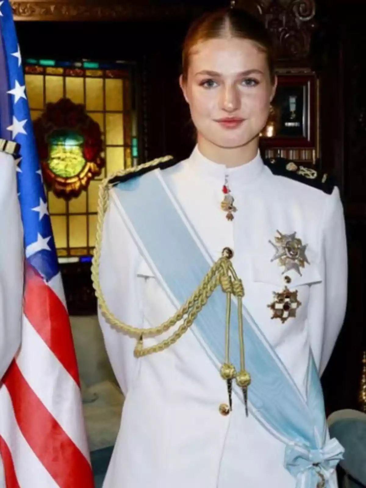Jove amb uniforme militar blanc amb xarreteres daurades i banda blau clar, dret al costat d’una bandera dels Estats Units en un entorn elegant Jove amb uniforme militar blanc amb xarreteres daurades i banda blau clar, dret al costat d’una bandera dels Estats Units en un entorn elegant