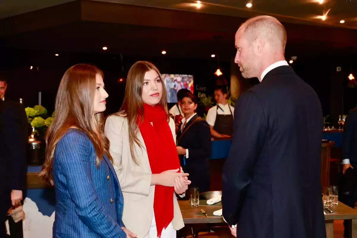 Leonor y Sofía conversan con el príncipe Guillermo en un ambiente elegante con personas al fondo Leonor y Sofía conversan con el príncipe Guillermo en un ambiente elegante con personas al fondo