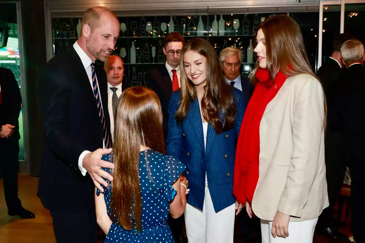 El príncipe Guillermo con traje saluda a Charlotre mientras Leonor y Sofía sonríen y observan la escena en un ambiente formal con varias personas al fondo. El príncipe Guillermo con traje saluda a Charlotre mientras Leonor y Sofía sonríen y observan la escena en un ambiente formal con varias personas al fondo.