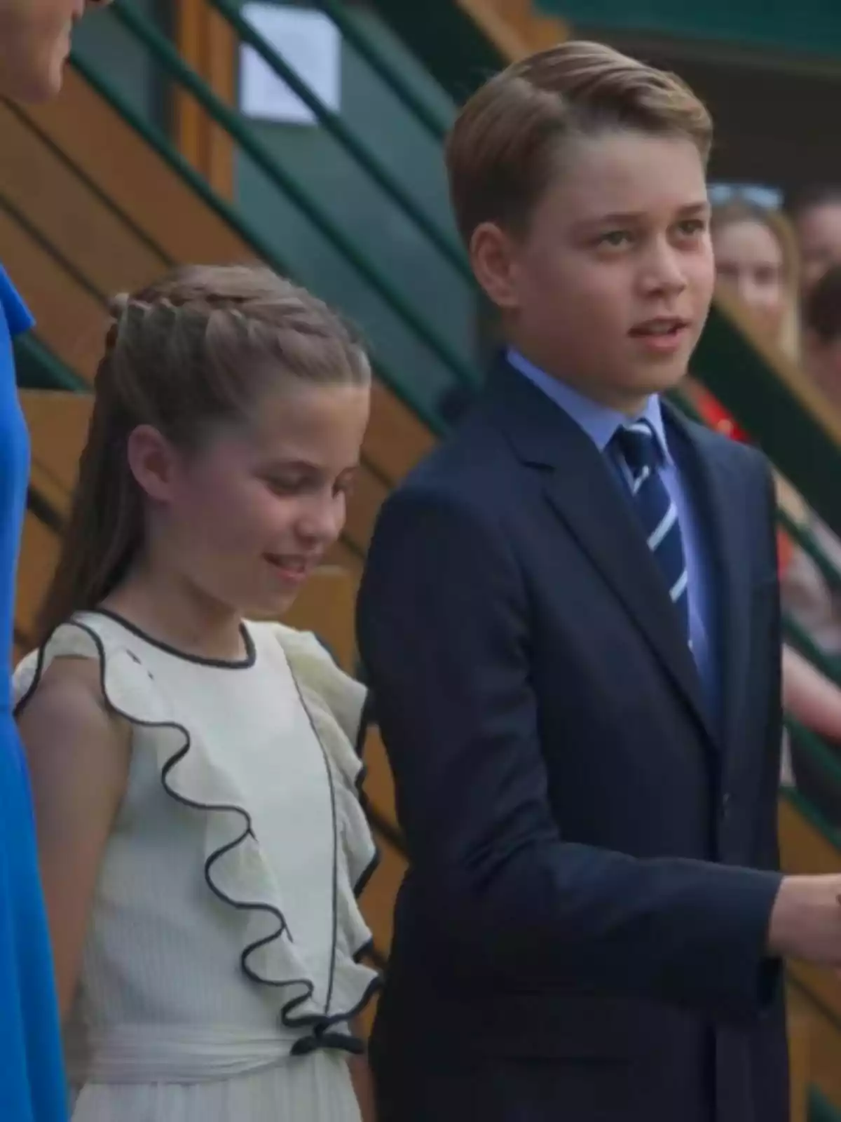 Two well-dressed children, one is Princess Charlotte in a white ruffled dress and the other is Prince George in a blue suit and tie, are standing together at a formal event.