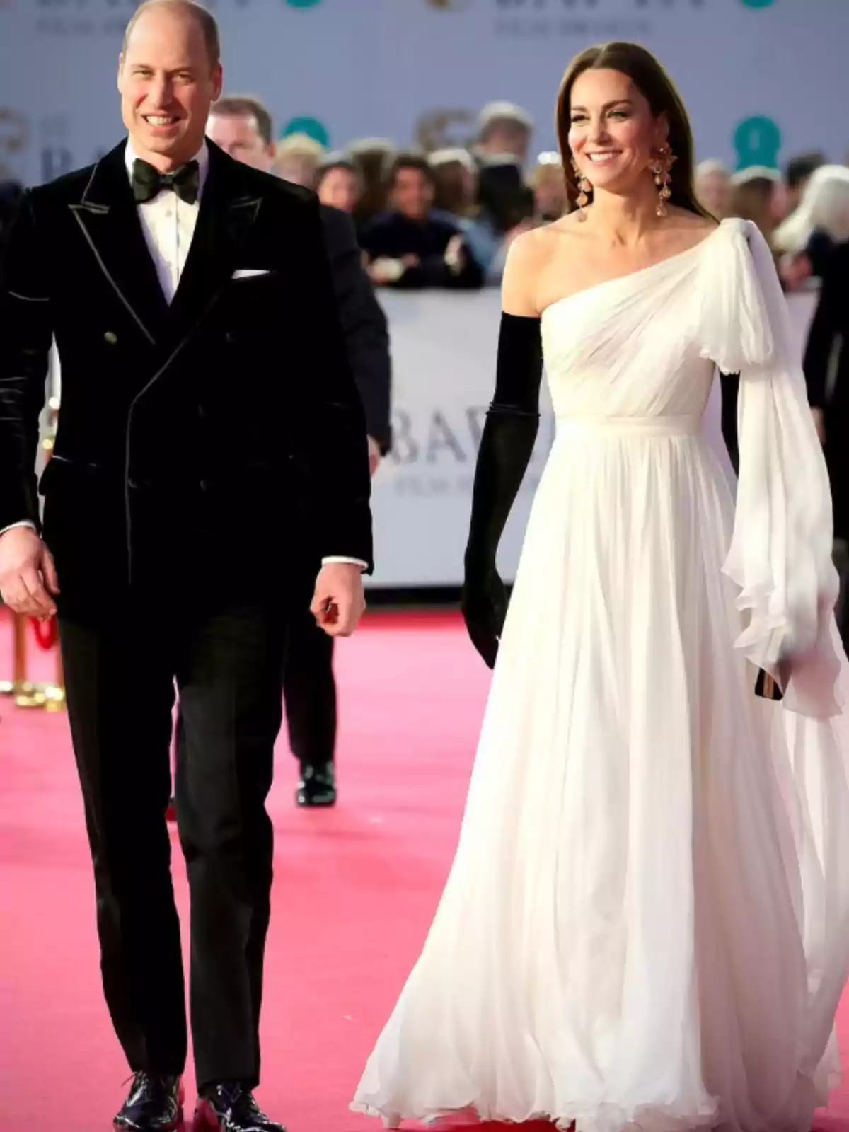 Pareja elegantemente vestida caminando por una alfombra roja, él con esmoquin negro y ella con vestido blanco de un solo hombro y guantes largos negros