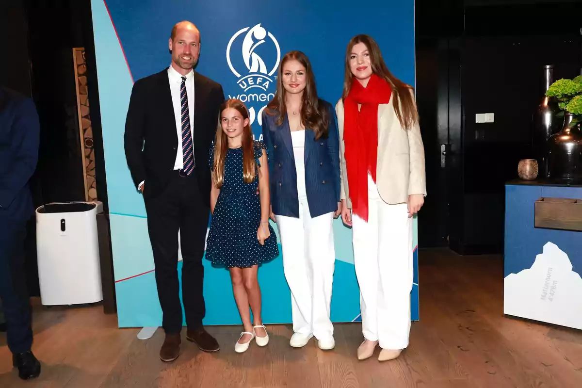 Leonor, Sofía, el príncipe Guillermo y su hija Charlotte posan sonrientes frente a un fondo azul con el logo de la UEFA Women’s Euro, dos de ellas llevan pantalón blanco y una bufanda roja, mientras que una niña lleva un vestido azul con lunares blancos. Leonor, Sofía, el príncipe Guillermo y su hija Charlotte posan sonrientes frente a un fondo azul con el logo de la UEFA Women’s Euro, dos de ellas llevan pantalón blanco y una bufanda roja, mientras que una niña lleva un vestido azul con lunares blancos.