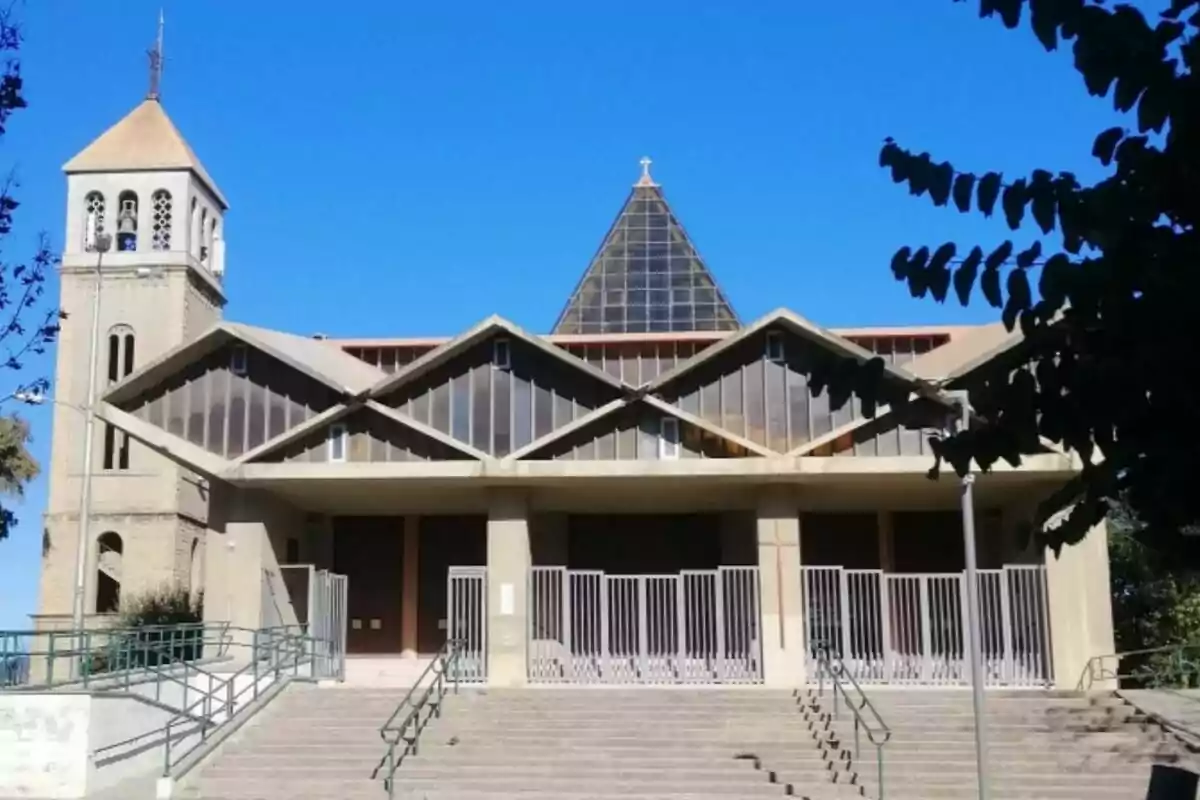 Façana d’una església moderna amb sostre en forma de zig-zag i un campanar a l’esquerra sota un cel serè Façana d’una església moderna amb sostre en forma de zig-zag i un campanar a l’esquerra sota un cel serè