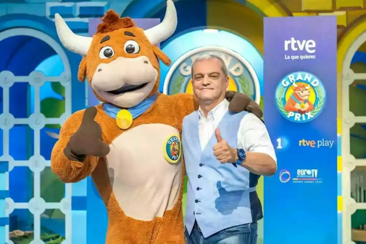 Un home somrient amb armilla blava posa al costat d’una mascota de toro en un plató de televisió colorit del programa Grand Prix. Un home somrient amb armilla blava posa al costat d’una mascota de toro en un plató de televisió colorit del programa Grand Prix.