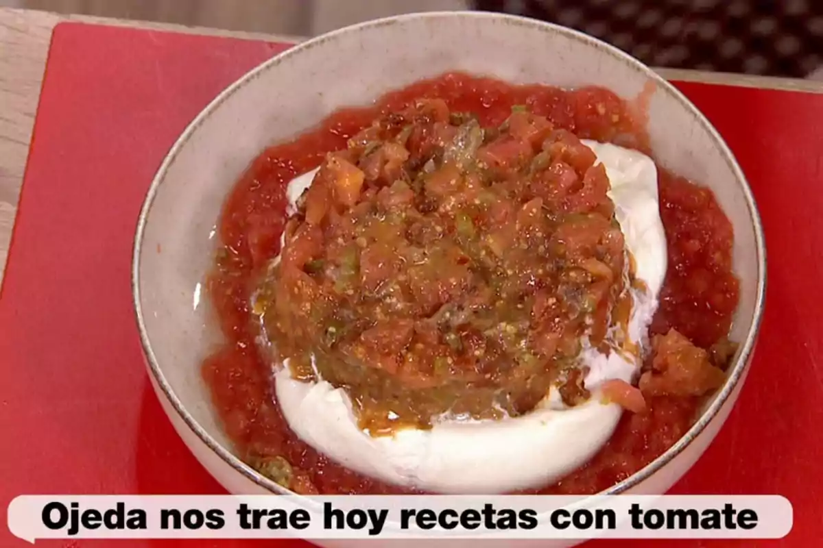 Plat fondo amb una base de salsa de tomàquet i una bola de formatge coberta amb una barreja de tomàquet picat, sobre una taula vermella i un text a la part inferior que diu Ojeda avui ens porta receptes amb tomàquet. Plat fondo amb una base de salsa de tomàquet i una bola de formatge coberta amb una barreja de tomàquet picat, sobre una taula vermella i un text a la part inferior que diu Ojeda avui ens porta receptes amb tomàquet.