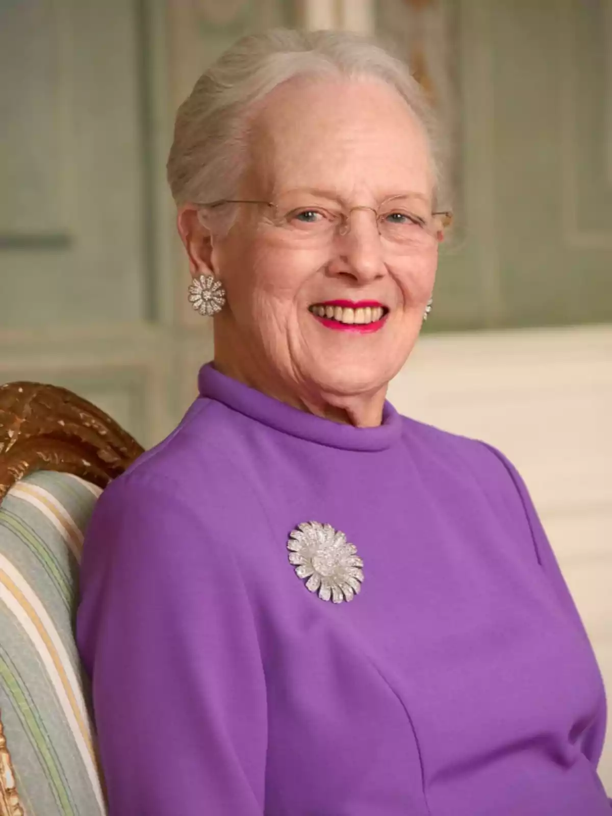 Elderly woman with gray hair and glasses sitting in an elegant chair, wearing a purple dress with a silver flower-shaped brooch and smiling at the camera. Elderly woman with gray hair and glasses sitting in an elegant chair, wearing a purple dress with a silver flower-shaped brooch and smiling at the camera.