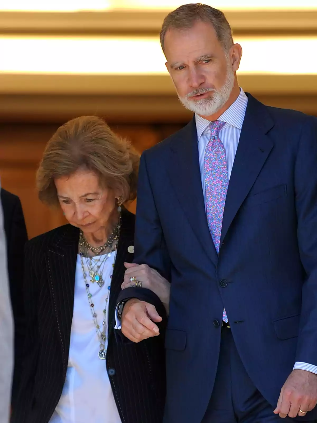 Felip VI amb vestit blau ajuda la reina Sofia, que porta un conjunt fosc i diversos collars. Felip VI amb vestit blau ajuda la reina Sofia, que porta un conjunt fosc i diversos collars.