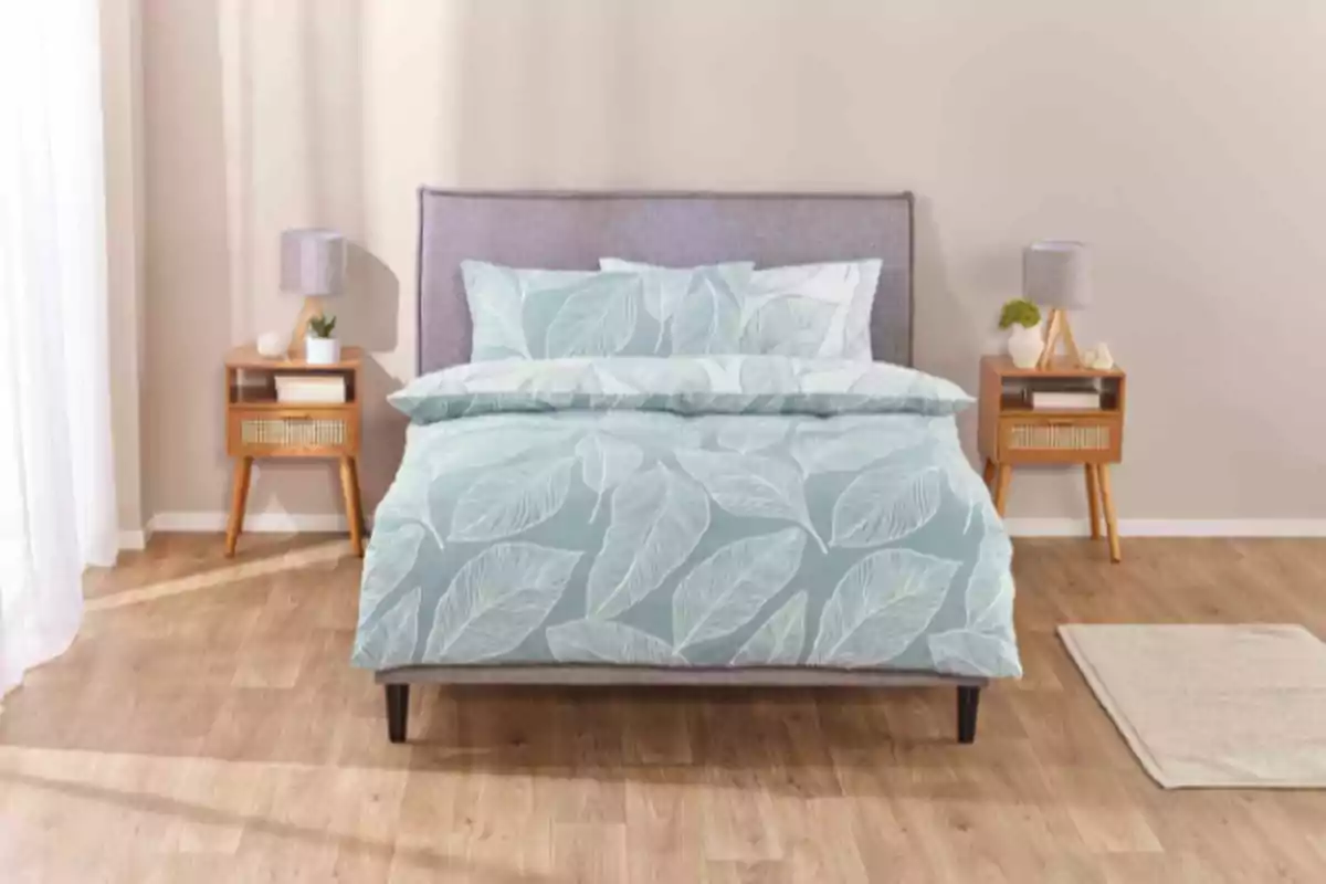 Llit amb edredó blau de fulles blanques en un dormitori modern amb dues tauletes de nit de fusta i làmpades grises Llit amb edredó blau de fulles blanques en un dormitori modern amb dues tauletes de nit de fusta i làmpades grises