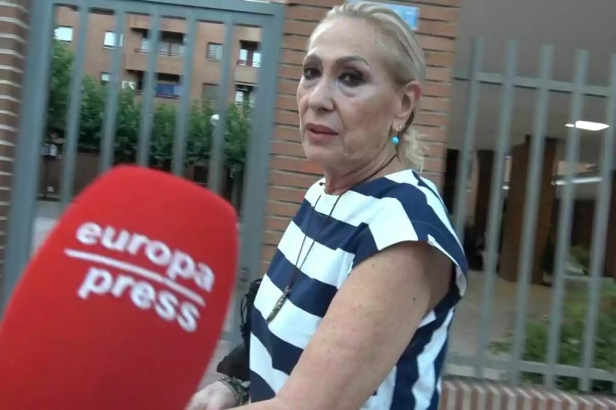 Rosa Benito amb samarreta de ratlles blanques i blaves sent entrevistada per un micròfon vermell d’Europa Press davant d’una reixa i edificis de maó. Rosa Benito amb samarreta de ratlles blanques i blaves sent entrevistada per un micròfon vermell d’Europa Press davant d’una reixa i edificis de maó.