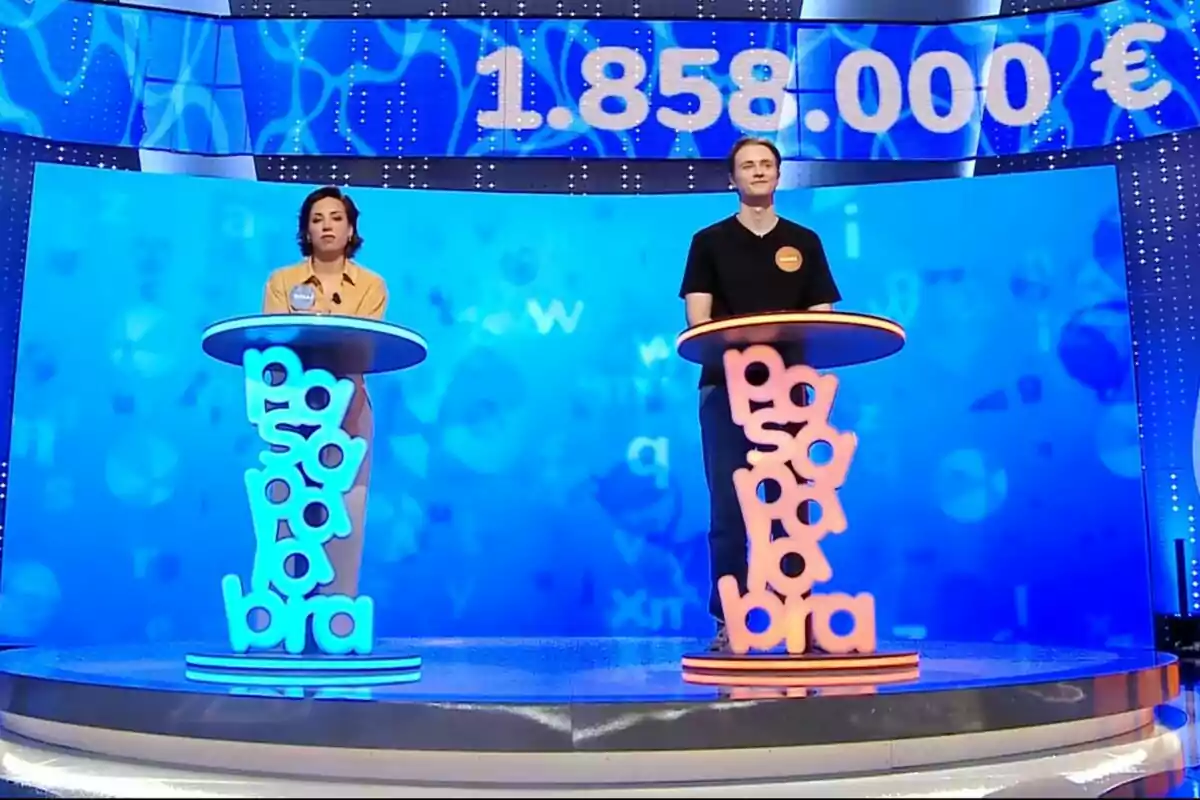 Dos concursants, que són la Rosa i en Manu, drets als seus respectius faristols al plató del programa de televisió 'Pasapalabra' amb un marcador gran que mostra un premi d’un milió vuit-cents cinquanta-vuit mil euros sobre un fons blau. Dos concursants, que són la Rosa i en Manu, drets als seus respectius faristols al plató del programa de televisió 'Pasapalabra' amb un marcador gran que mostra un premi d’un milió vuit-cents cinquanta-vuit mil euros sobre un fons blau.