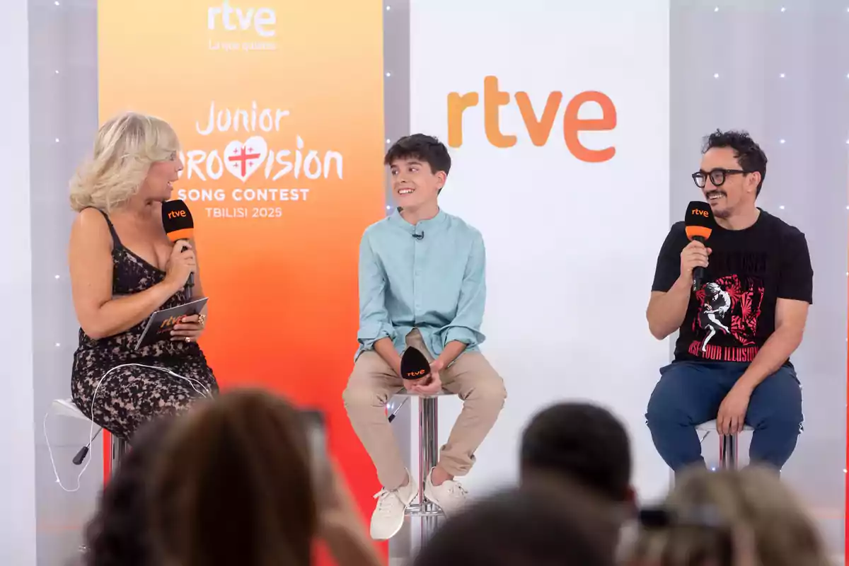 Tres persones assegudes en tamborets conversen i somriuen durant una presentació de premsa de RTVE per al Junior Eurovision Song Contest 2025 en un escenari amb fons taronja i blanc. Tres persones assegudes en tamborets conversen i somriuen durant una presentació de premsa de RTVE per al Junior Eurovision Song Contest 2025 en un escenari amb fons taronja i blanc.