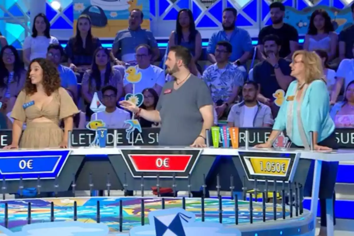 Tres concursantes participan en un programa de juegos de televisión mientras el público observa desde las gradas Tres concursantes participan en un programa de juegos de televisión mientras el público observa desde las gradas