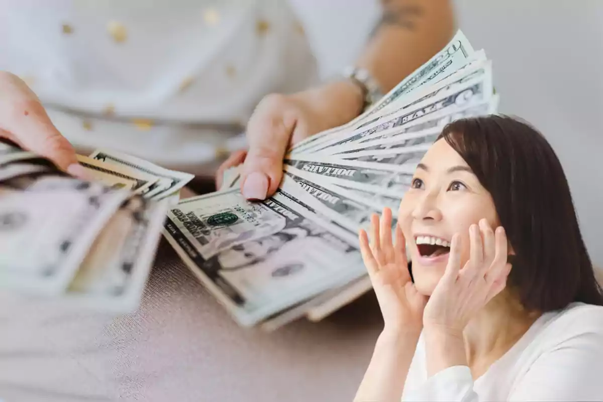 Una mujer sonriente y sorprendida observa un fajo de billetes de dólar sostenidos por otra persona.