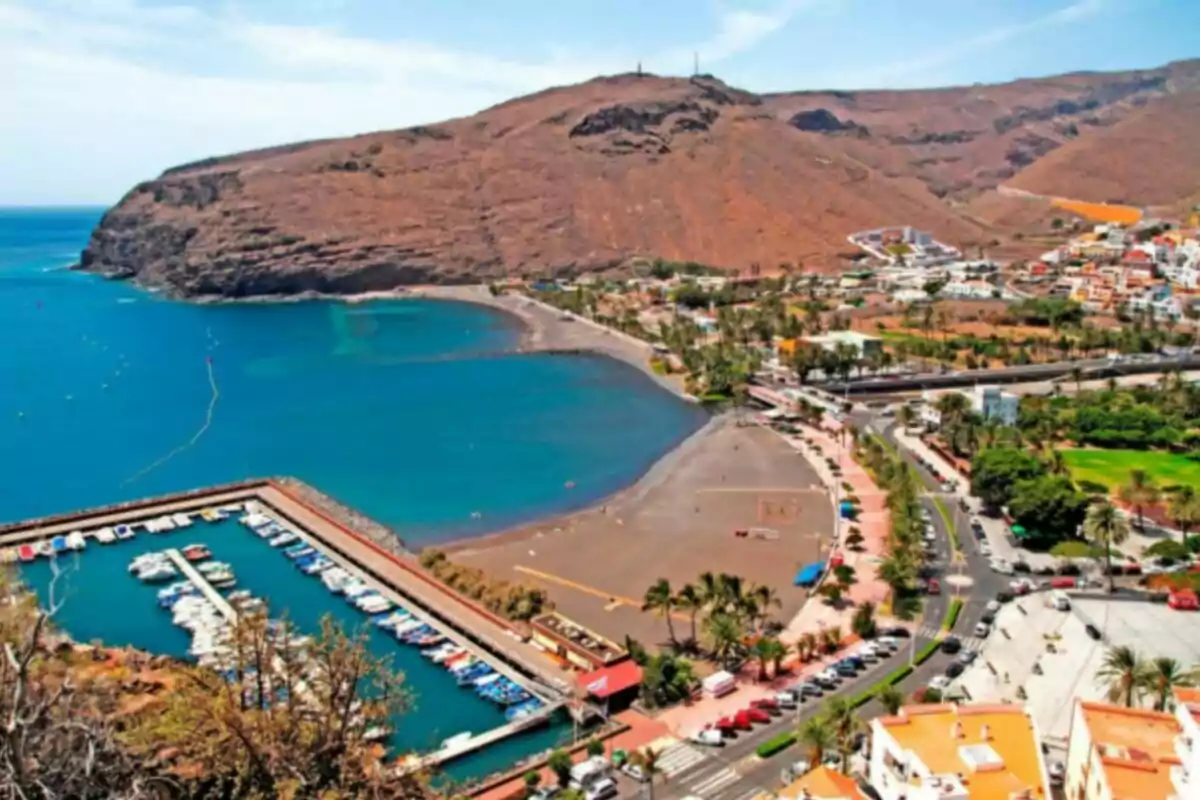 Vista panorámica de una bahía con playa de arena oscura, puerto deportivo con barcos y un pueblo costero rodeado de montañas áridas Vista panorámica de una bahía con playa de arena oscura, puerto deportivo con barcos y un pueblo costero rodeado de montañas áridas