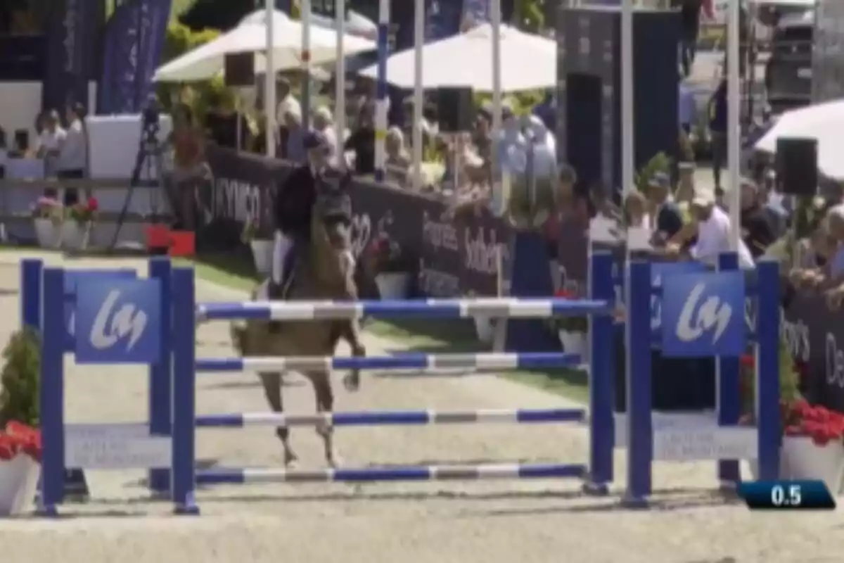 A rider on horseback, Sira Martínez, prepares to jump an obstacle in an outdoor equestrian competition, with spectators and judges watching from the background.
