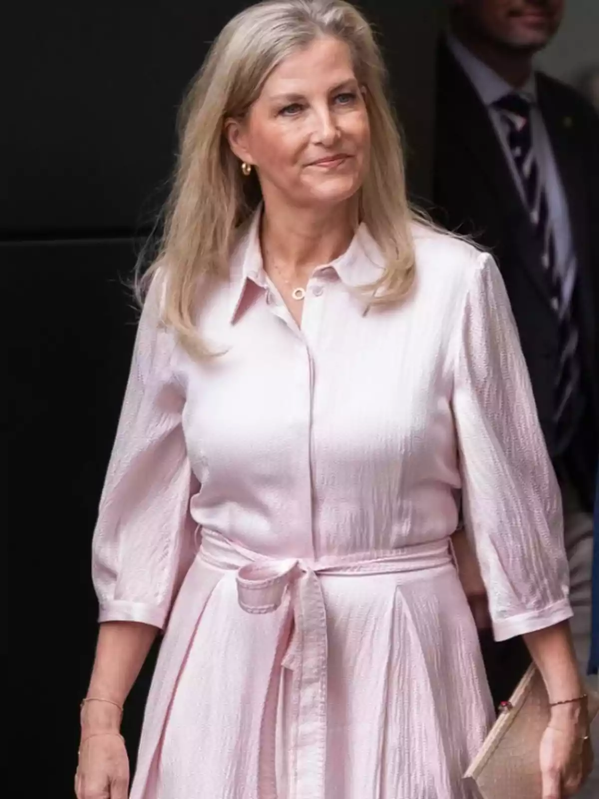 Dona de cabell ros i llarg vestida amb un vestit rosa clar de màniga tres quarts i cinturó, sostenint una bossa beix i somrient lleument. Dona de cabell ros i llarg vestida amb un vestit rosa clar de màniga tres quarts i cinturó, sostenint una bossa beix i somrient lleument.