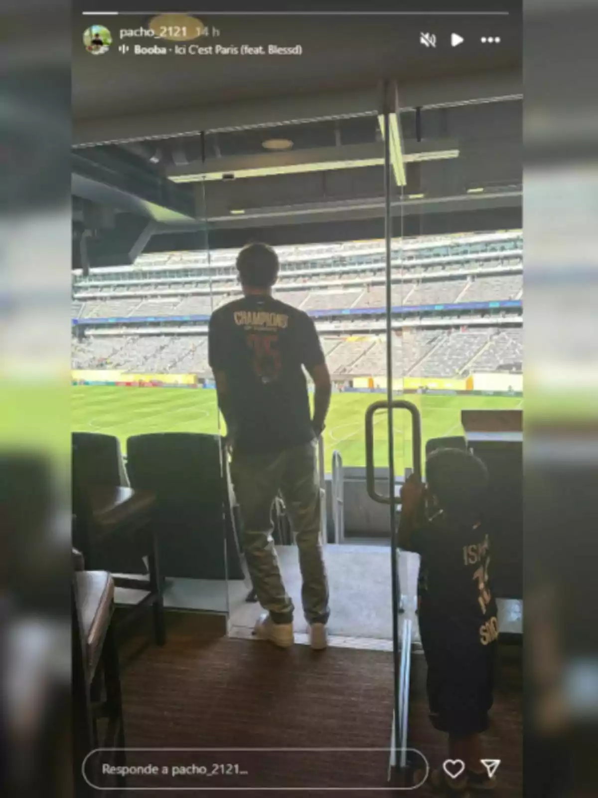 Pacho Martínez y un niño de espaldas observan un estadio de fútbol desde un palco, ambos llevan camisetas oscuras con letras en la espalda Pacho Martínez y un niño de espaldas observan un estadio de fútbol desde un palco, ambos llevan camisetas oscuras con letras en la espalda